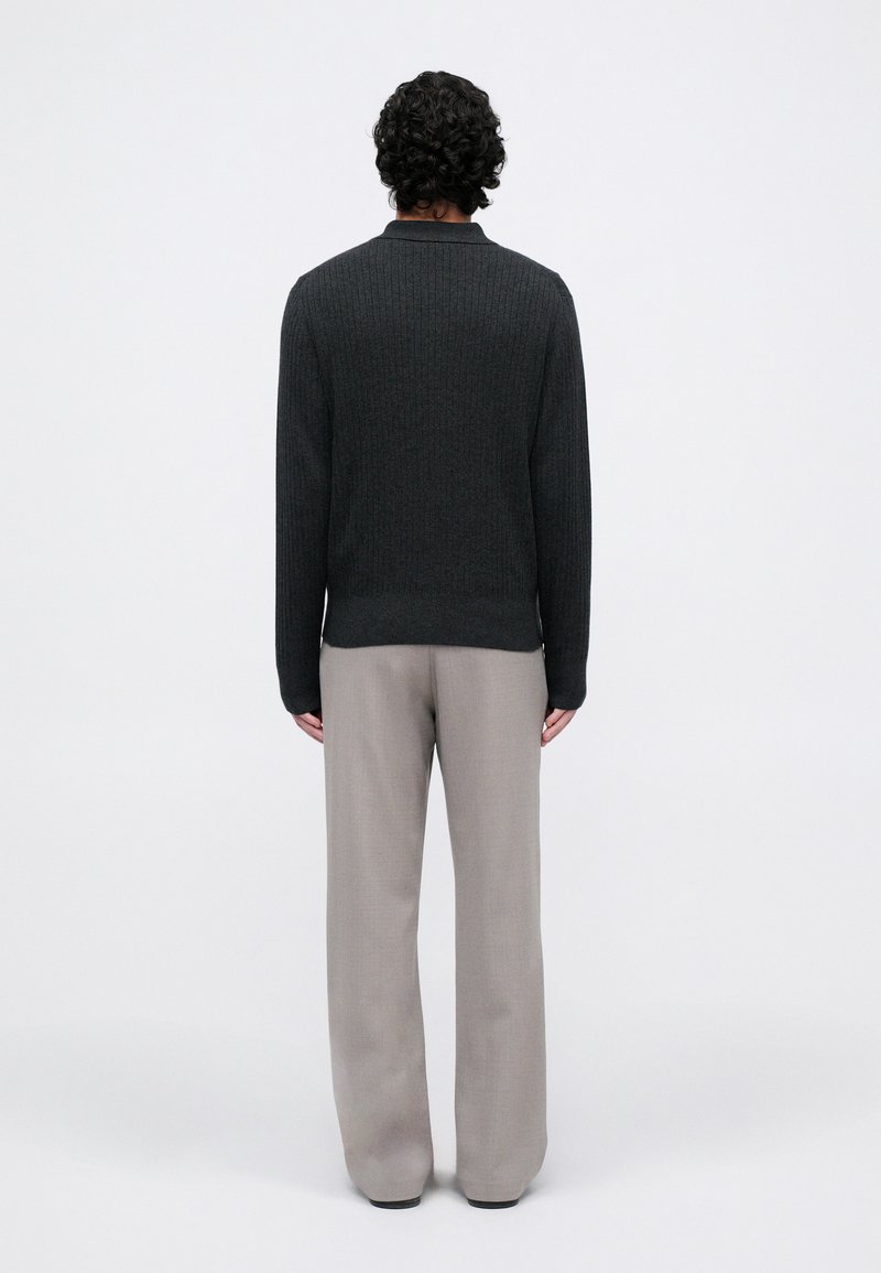 Man standing facing away, wearing a black ribbed sweater and loose-fitting light gray pants against a plain white background.