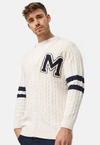 Creme kabelstrikket cardigan med en marineblå "M" lap og stribede accenter på ærmerne. Designet med V-udskæring og knaplukning.