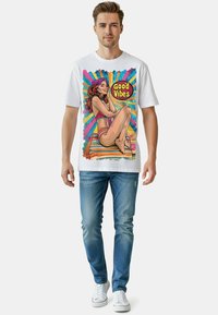 Weißes T-Shirt mit einem bunten Grafikbild einer Frau auf einem gestreiften Hintergrund mit dem Text "Good Vibes". Kombiniert mit blauen Jeans und weißen Sneakers.
