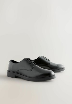 Zapatos de cuero negro con cordones, puntera redonda, superficie lisa y suela plana. Presentan un sencillo pespunte a lo largo de los costados.