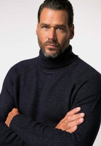 Maglione a dolcevita blu navy realizzato in un tessuto a maglia testurizzato, con un design aderente e polsini a coste, indossato da una persona con le braccia incrociate.