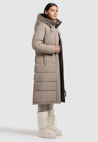 Lange beige gewatteerde jas met een hoge kraag, horizontaal gestikt patroon, ritssluiting aan de voorkant en zijzakken. Gecombineerd met witte sneeuwlaarzen.