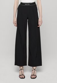 Svarte vidbukse med høyt liv og hvite sømmer. Glatt tekstur og rene linjer, kombinert med svarte sandaler med stropper.