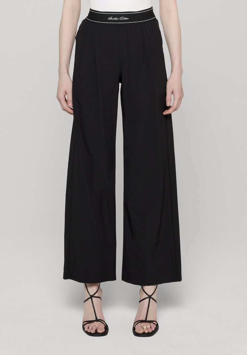 Svarte vidbukse med høyt liv og hvite sømmer. Glatt tekstur og rene linjer, kombinert med svarte sandaler med stropper.