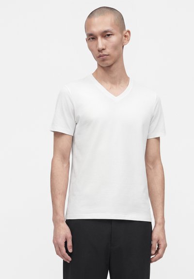 Homme aux cheveux courts portant un t-shirt blanc uni à manches courtes avec col en V et un pantalon noir, debout devant un fond clair.