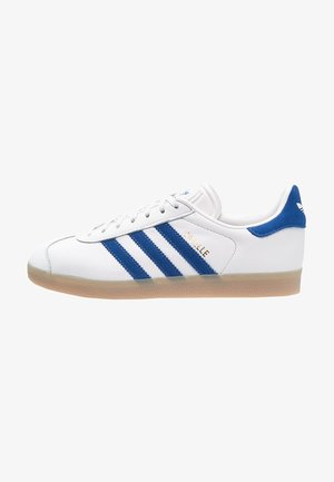 Białe niskie sneakersy Adidas z niebieskimi paskami, niebieską pętelką na pięcie, gumową podeszwą i złotym napisem "Gazelle" na bocznym panelu.
