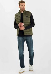 Chaleco acolchado verde oliva con diseño acolchado, cuello alto y cierre de cremallera, usado sobre una camiseta negra de manga larga y vaqueros azules. Zapatillas blancas.