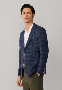 Muž v modrom károvanom saku, bielej košeli a olivovo zelených nohaviciach stojaci s jednou rukou v vrecku na jednoduchom pozadí.