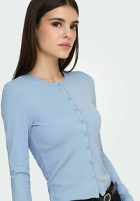 Haut côtelé bleu clair à manches longues avec col rond, fermeture boutonnée sur le devant et détail de ourlet brut. Texture lisse, coupe ajustée.