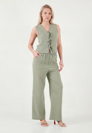 Femme portant un haut sans manches vert clair à nouer sur le devant et un pantalon large assorti, debout avec une main dans la poche et des talons colorés.