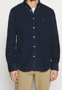 Camicia di velluto a coste blu navy con colletto button-down, maniche lunghe e tasca frontale. Presenta bottoni bianchi e un logo discreto sulla tasca del petto.