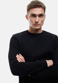 Sudadera negra de cuello redondo hecha de una tela suave, con puños y cuello de canalé. El modelo tiene los brazos cruzados, mostrando una silueta ajustada.