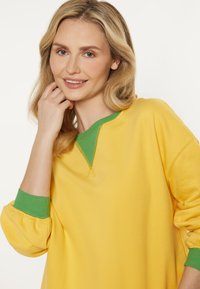 Geel sweatshirt met groene accenten op de kraag en manchetten, met een relaxte pasvorm en een zachte textuur. Ook zichtbaar stikseldetail.