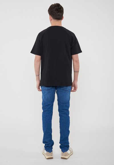 T-shirt nera in cotone a maniche corte, con scollatura tonda e spalle raglan, abbinata a jeans in denim blu e sneakers beige.