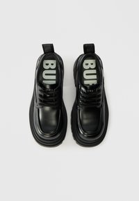 Melnas ādas kurpes ar apaļiem purngaliem, biezu zoli un auklām. Iekšpusē ir pelēks iebūvētais zole ar treknu melnu "BUFF" tekstu; visā virsmas apstrādē ir spīdīgs finish.