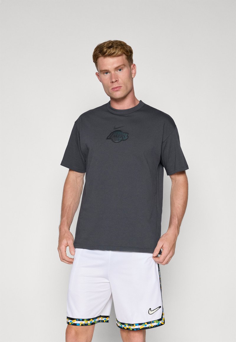 Grå t-shirt med korte ærmer og en sort 'Lakers' logo, sammen med hvide basketballshorts med en farverig mønstret kant og Nike-logo.