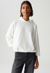 Vrouw in een witte getextureerde hoodie en zwarte jeans, staand met één hand op haar heup tegen een eenvoudige lichte achtergrond.
