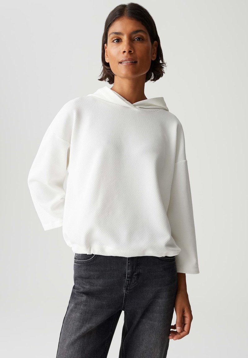 Vrouw in een witte getextureerde hoodie en zwarte jeans, staand met één hand op haar heup tegen een eenvoudige lichte achtergrond.
