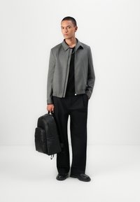 Jachetă bomber gri cu guler, tricou negru, pantaloni negri largi și un rucsac din piele neagră texturată cu multiple compartimente.