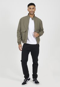 Oliwkowa kurtka bomberka z stójką, dwiema przednimi kieszeniami, zapinana na zamek, połączona z białą koszulką, czarnymi spodniami i czarnymi sneakersami.