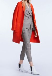 Manteau en laine orange, blazer croisé à carreaux gris, pantalon ajusté à carreaux gris et bottines blanches avec accents noirs.