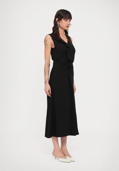 Sieviete ar tetovējumiem valkā melnu midi kleitu bez piedurknēm ar volānu detaļu, ko papildina baltas smailas kāju papēžu čības ar zemu papēdi un garas auskari.