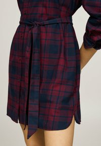 Rutig skjortekjole i rød og marineblå, med bæltestrop i taljen, knaplukning foran og sideslids. Lavet af blødt, struktureret stof.