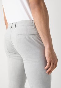 Gros plan d’une personne portant un pantalon gris clair ajusté et une chemise blanche, le bras droit détendu le long du corps.