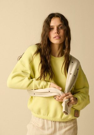 Jonge vrouw met lang bruin haar die een lichtgele trui draagt en een beige trui met kleine bruine hartjes over haar schouder houdt.