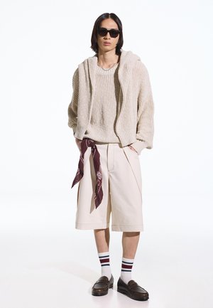 Mand iført beigefarvet striktrøje og shorts, mørke solbriller, stribede sokker, brune loafers og en vinrød bandana, der hænger fra lommen.