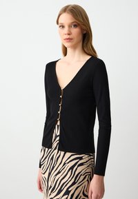 Zwarte geribbelde cardigan met een diepe V-hals en gouden knoopaccenten in het midden. Lange mouwen en een getailleerd ontwerp.