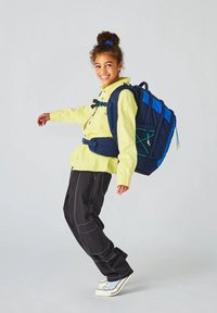 Rucksack aus marineblauem Stoff, mit mehreren Fächern und blauen Akzenten. Verfügt über verstellbare Riemen und grüne Gummibänder. Modell trägt ein hellgelbes Shirt und schwarze Hosen.