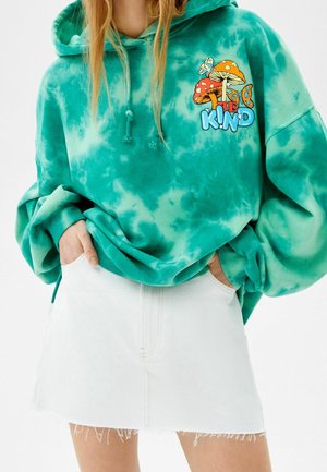 Felpa tie-dye in tonalità verde acqua con una grafica che presenta funghi e il testo "KIND." Abbinata a una gonna di jeans bianca con orlo sfilacciato.