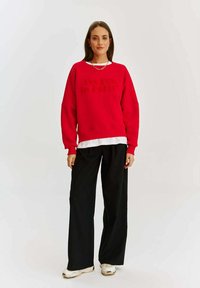 Červená mikina s vyrazeným textom "Uvidíme sa v Paríži" nosená cez bielu vrstvenú košeľu, v kombinácii s voľnými čiernymi nohavicami a bielymi teniskami.