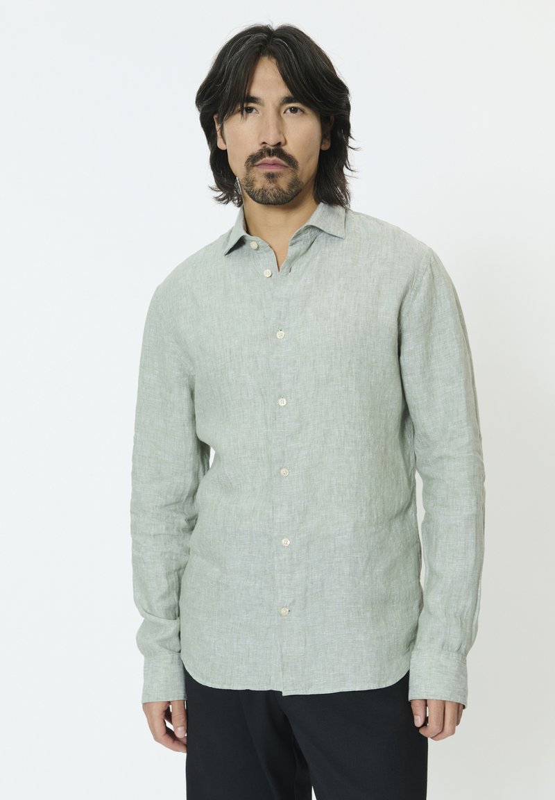 Homme aux cheveux mi-longs foncés et à la barbe, portant une chemise à manches longues vert clair boutonnée et un pantalon noir, debout devant un fond blanc.
