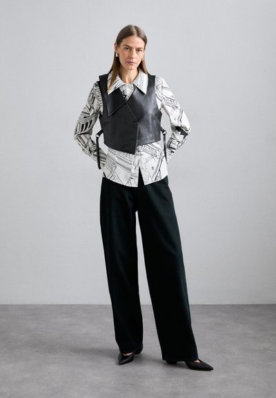 Chaleco de cuero negro sobre una camisa blanca con patrones abstractos en negro; combinado con pantalones de pierna ancha oscuros y zapatos negros puntiagudos.