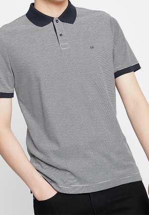 Homme portant un polo à manches courtes rayé gris et blanc avec col et poignets noirs, une main dans la poche du pantalon noir.
