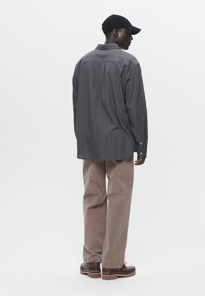 Graues Langarmshirt mit vertikalen Streifen, braune Cordhosen und braune Schnürschuhe, von hinten betrachtet. Schwarze Mütze getragen.