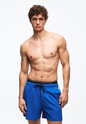 Mladý muž kučeravými vlasmi v modrých plaveckých šortkách stojaci pred svetlým jednoduchým pozadím, pozerajúci sa dopredu s neutrálnym výrazom.