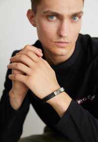 Black braided leather bracelet with a silver rectangular clasp featuring a logo, worn on a model's wrist with hands clasped.