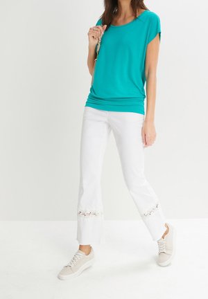 Kvinde iført teal top med korte ærmer, hvide bukser med blonde detaljer nær kanten og beige sneakers, stående mod en ensartet lys baggrund.