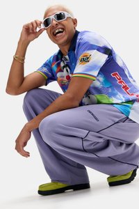 Camisa colorida com colarinho e gráfico de cartoon, combinada com calças largas lilás e sapatos verdes. Modelo usa óculos de sol brancos com detalhes em dourado.