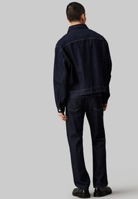 Combinaison en denim lavé foncé, comprenant une coupe décontractée, des coutures apparentes et des manches longues avec des poignets à boutons.
