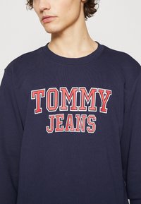 Sudadera azul marino de algodón, con un destacado logo en rojo y blanco "TOMMY JEANS" en el pecho. Cuello redondo, puños acanalados.