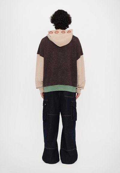 Sweatshirt surdimensionné à capuche avec un design en blocs de couleurs marron foncé et beige, ourlet vert et accents à motifs rouges sur la capuche ; associé à un pantalon cargo à jambes larges.