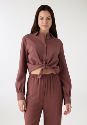 Camicia rosa cipria corta con bottoni e maniche lunghe, annodata sul davanti. Abbinata a pantaloni coordinati, ampi e morbidi. Texture liscia.