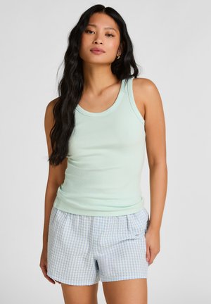 Femme aux longs cheveux noirs portant un haut vert clair sans manches et un short à carreaux bleu et blanc, debout avec une main dans la poche.