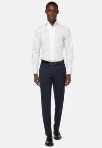 Weißes Hemd mit steifem Kragen, kombiniert mit navyblauen Hosen und schwarzen Lederschuhen, vervollständigt ein formelles Outfit.