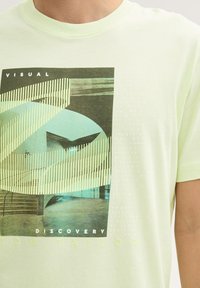 Lichtgroene katoenen t-shirt met een grafische print van abstracte vormen en tekst. Het ontwerp bevat strepen en accenten in blauw en zwart.