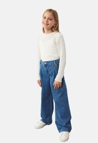 Witte, textuurrijke lange mouw top in combinatie met blauwe, wijdbenige denim jeans en witte sneakers. Eenvoudig ontwerp met een ontspannen pasvorm.